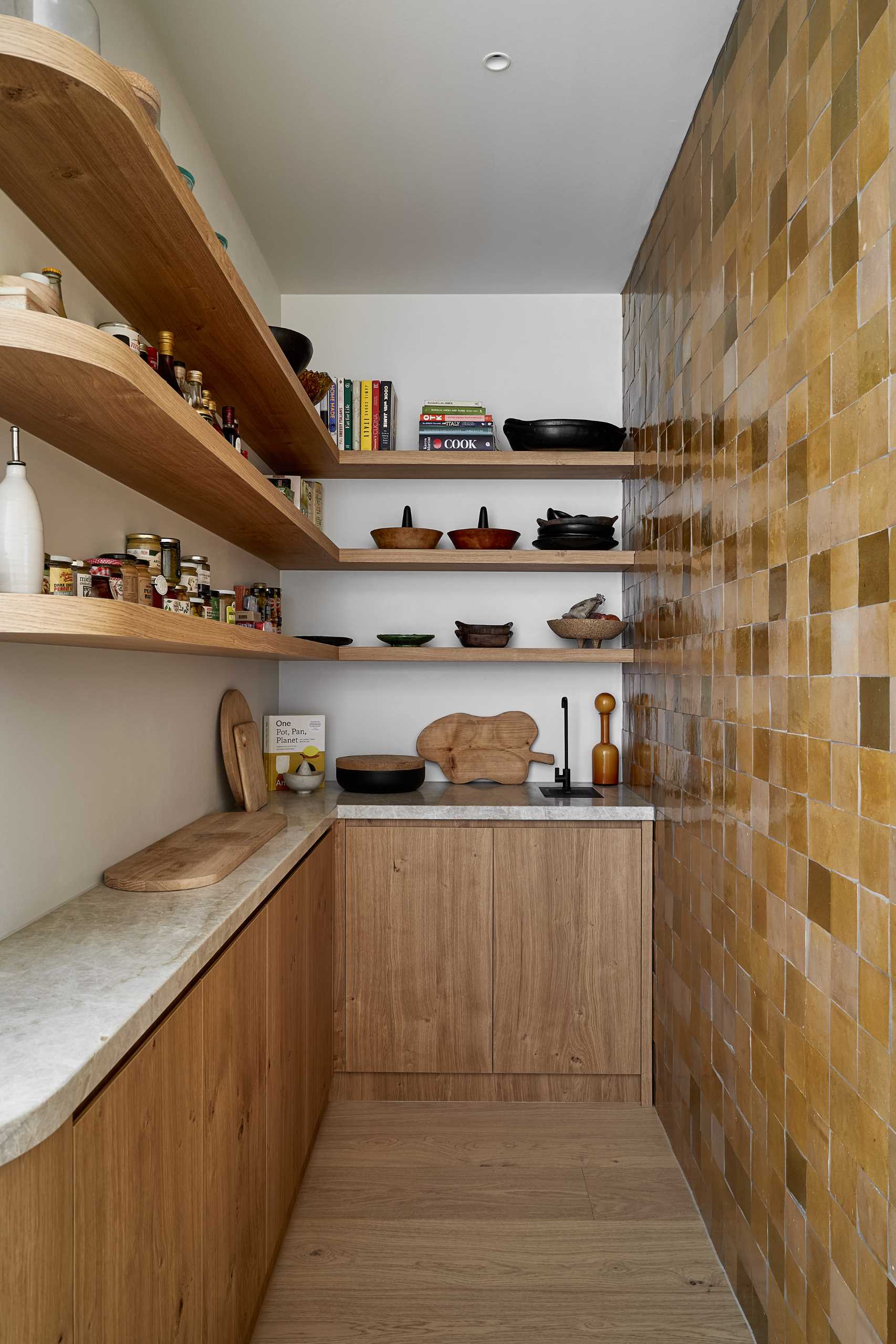 The kitchen is located at the end of the open-plan interior, with a tiled wall creating an eye-catching accent wall. Hidden from view is a pantry with additional storage and floating shelves.